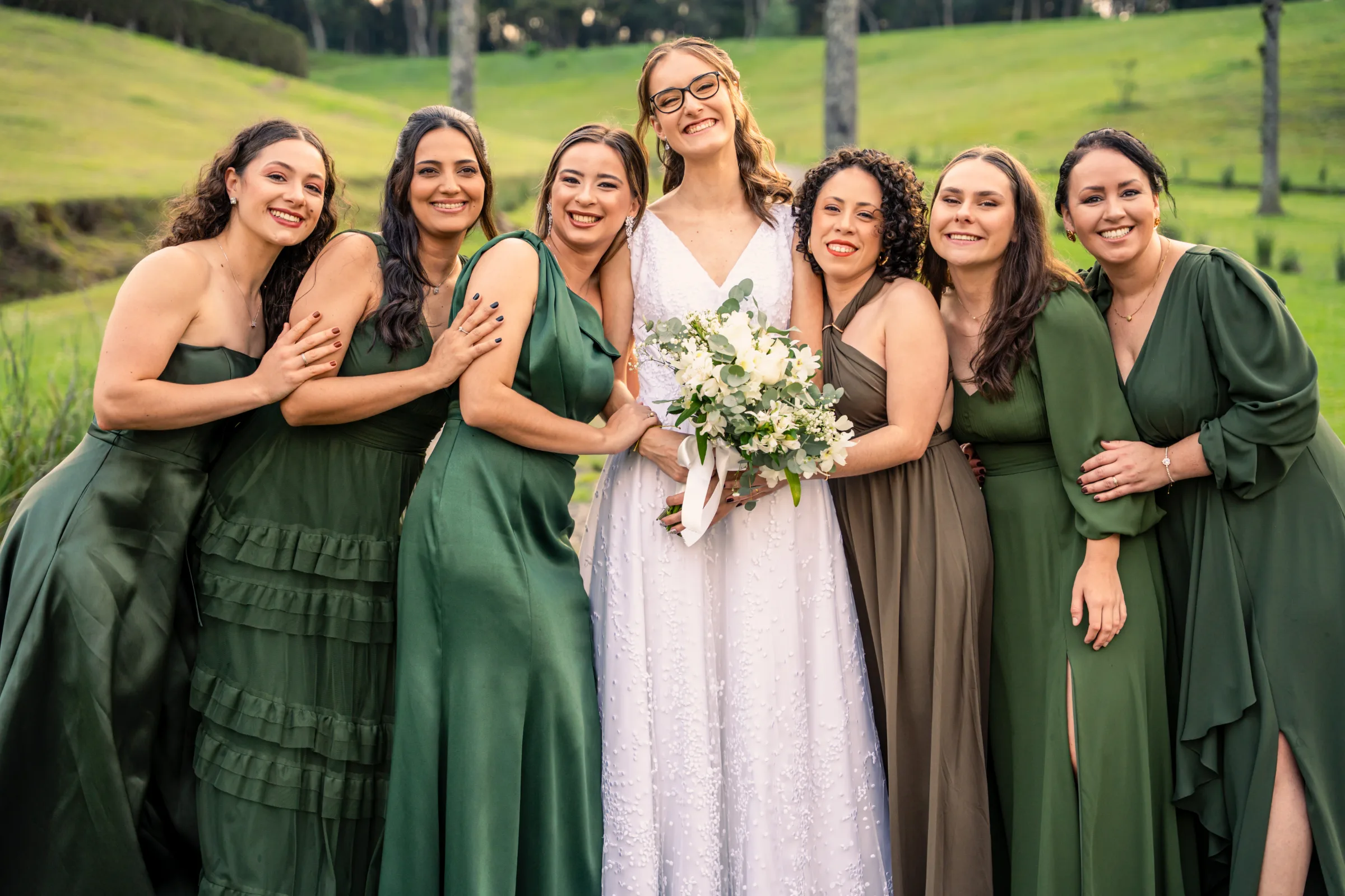 Noiva com buquê branco entre seis madrinhas de vestidos verdes no campo
