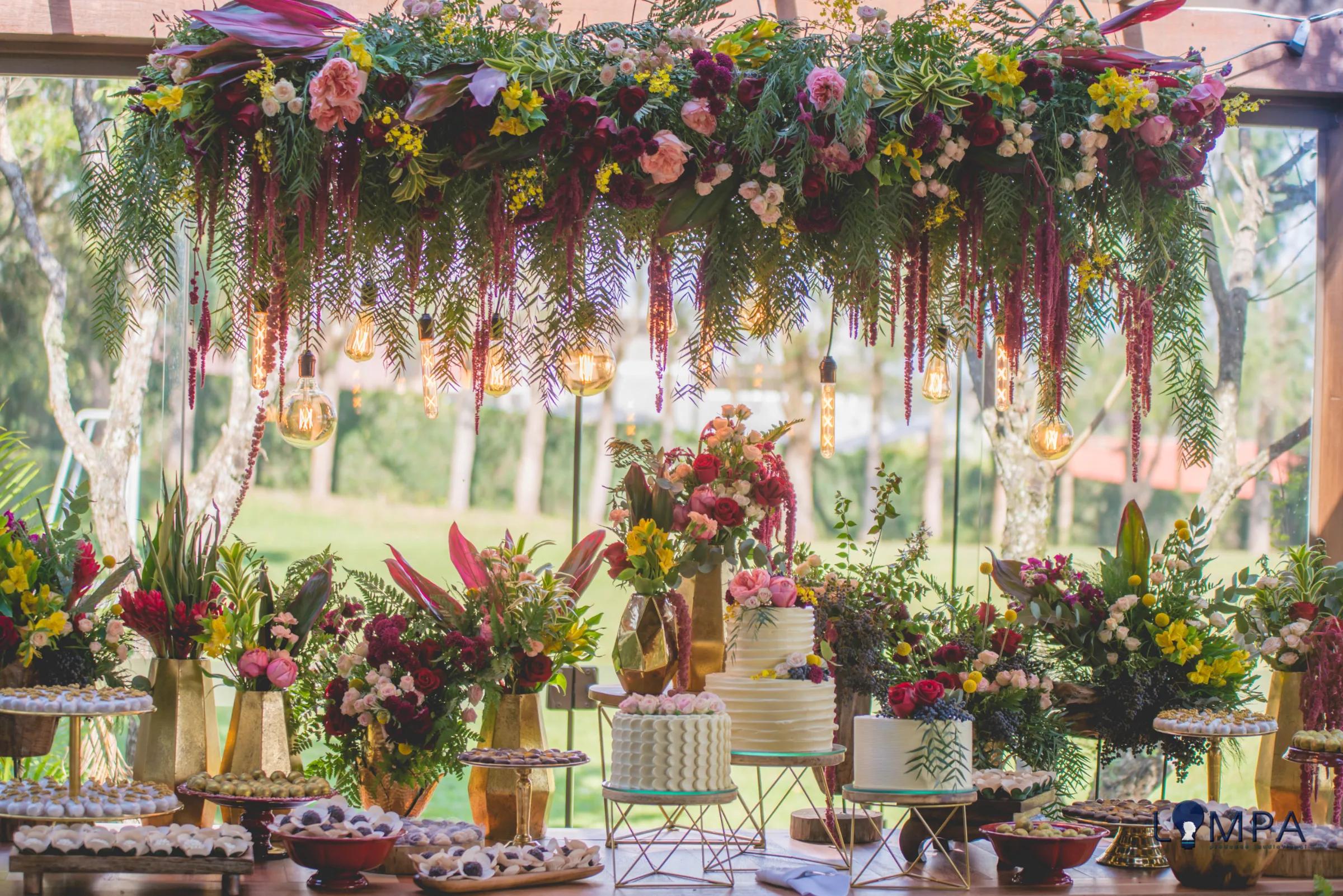 Bolo e doces sob dossel de flores com lâmpadas pendentes diante do vidro