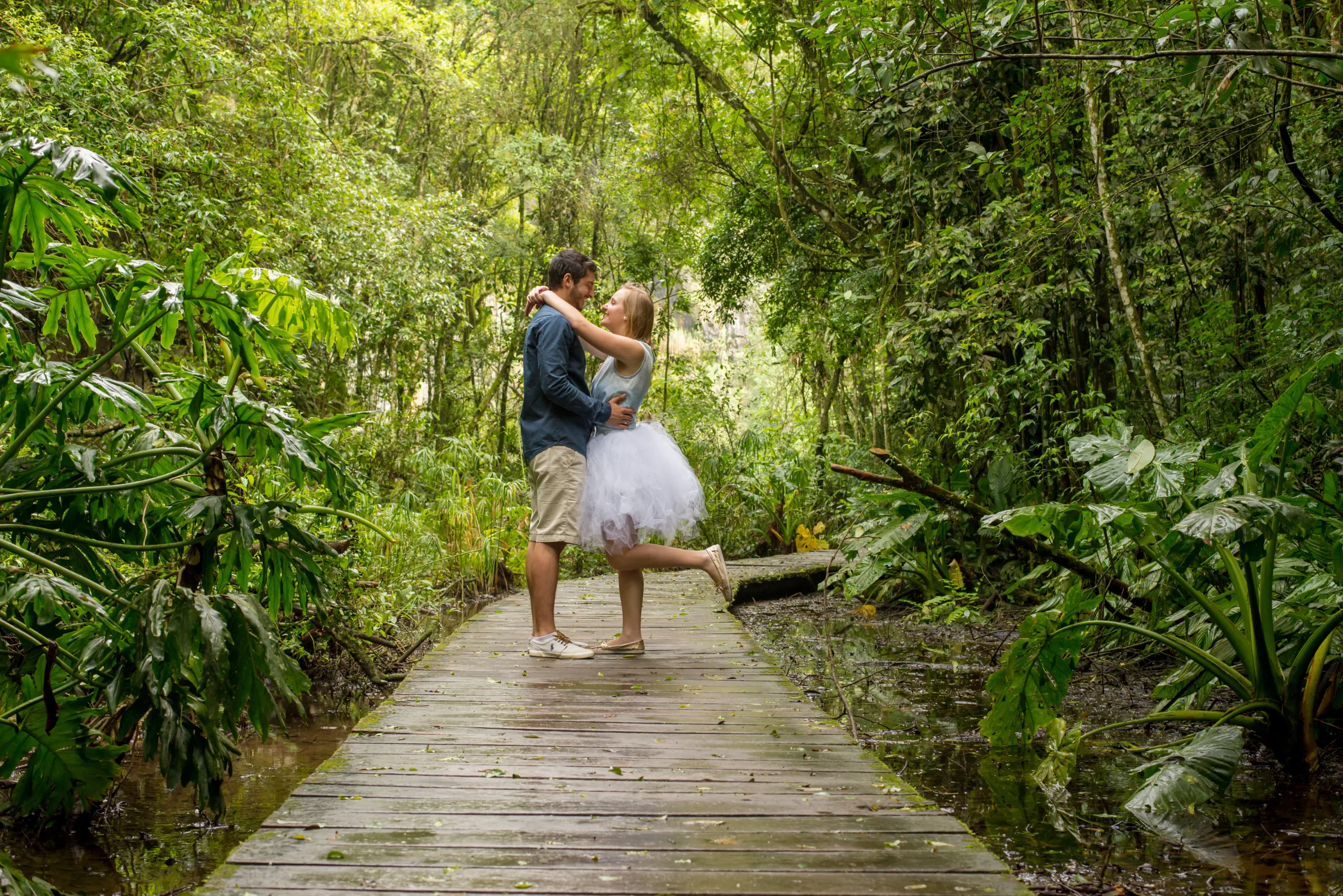 Casal abraçado em passarela de madeira cercada pela mata fechada
