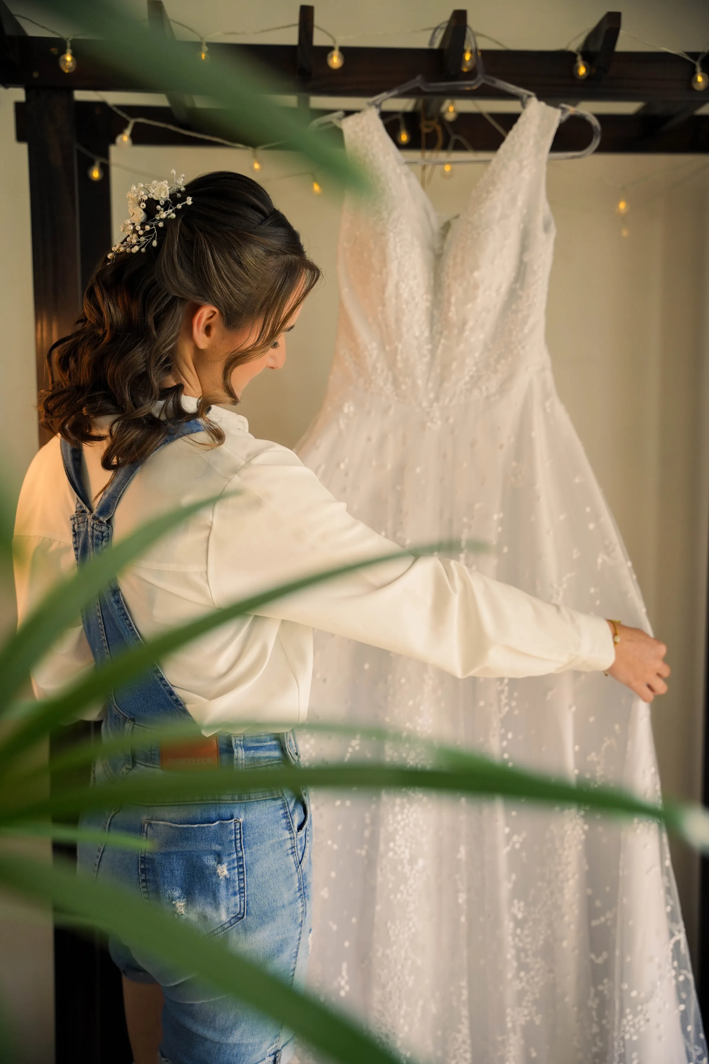 Noiva de macacão jeans toca o vestido pendurado sob fio de luzinhas