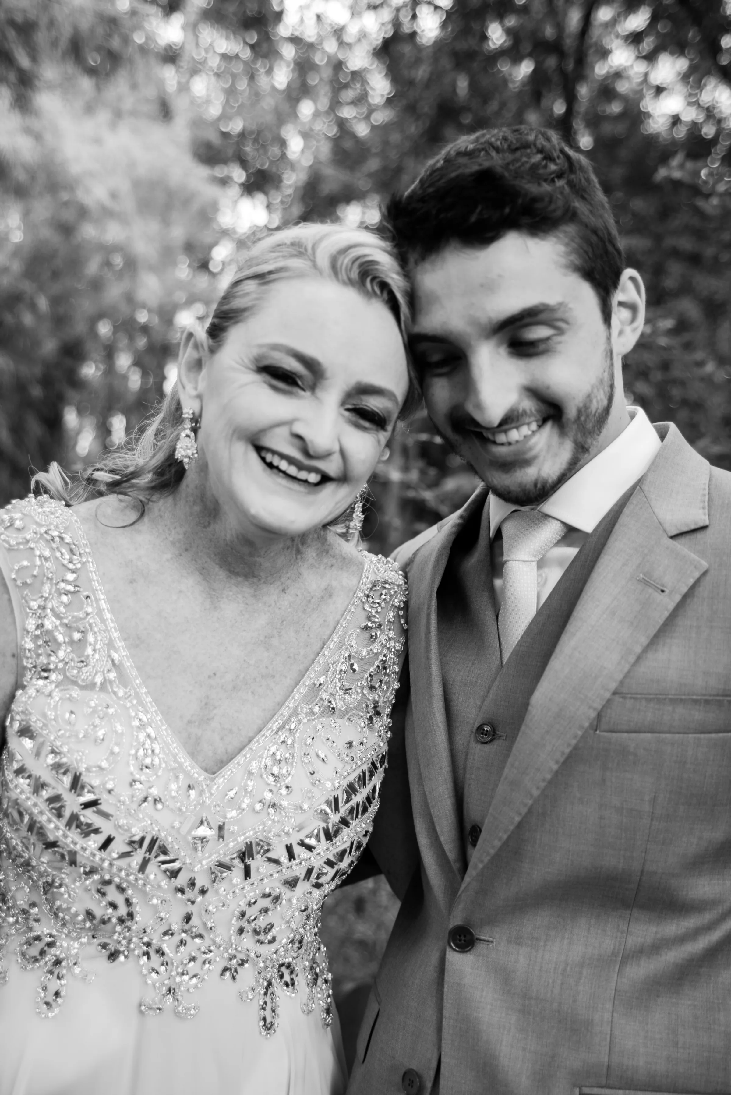 Rapaz de terno e mulher de vestido bordado rindo juntos, preto e branco