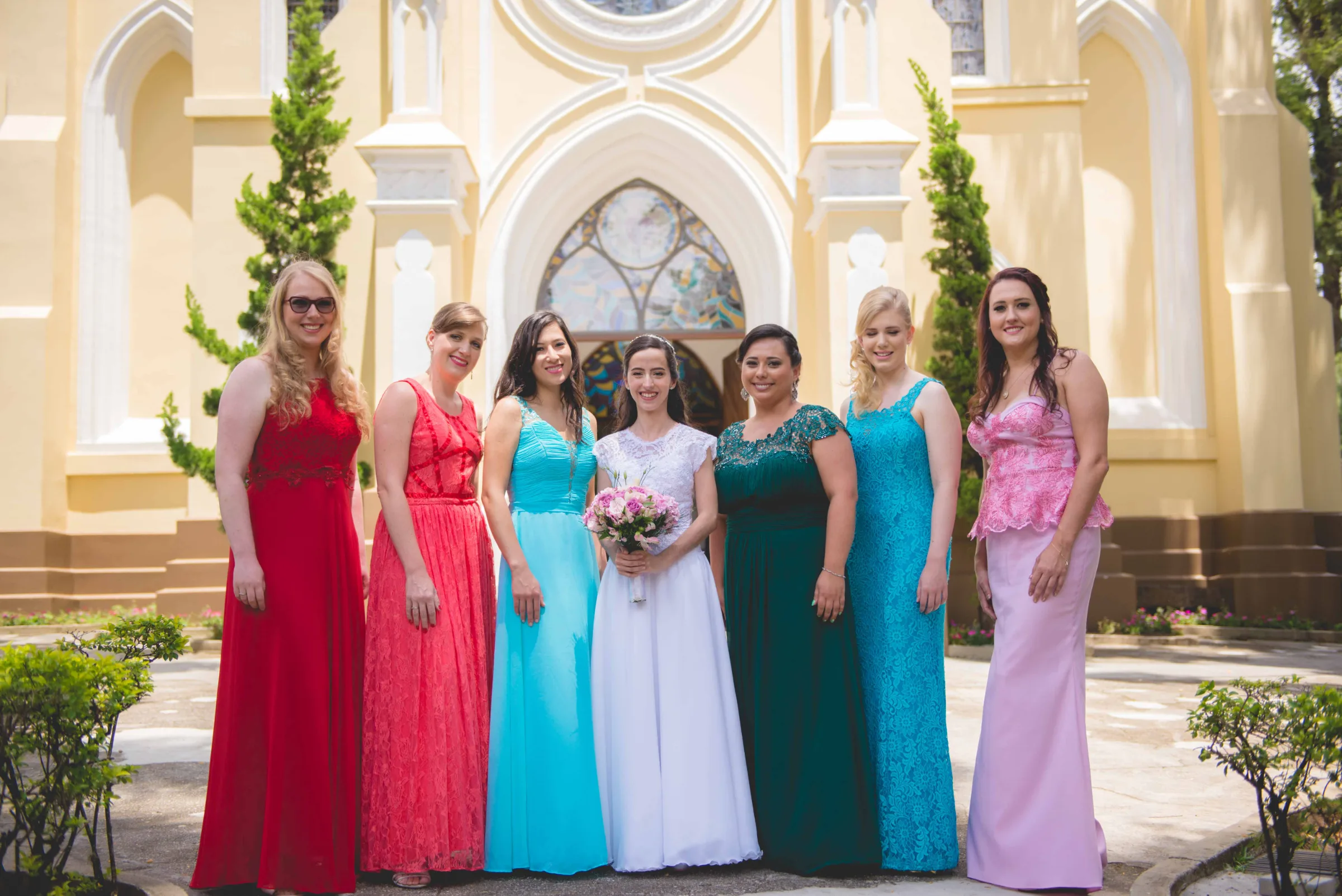 Noiva entre seis madrinhas de vestidos coloridos na escadaria da igreja amarela