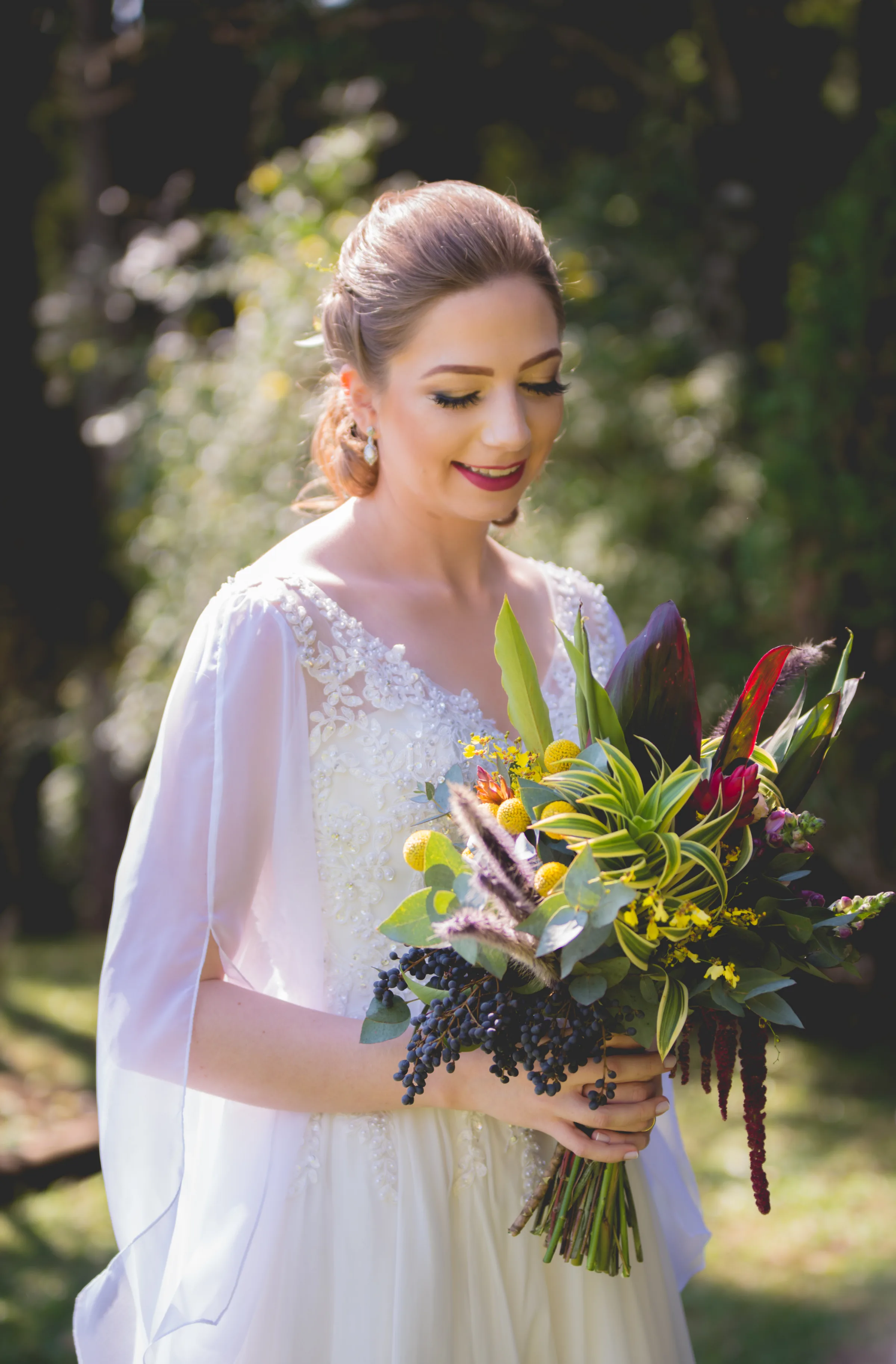 Noiva de olhar baixo segurando buquê de folhagens e flores amarelas no jardim