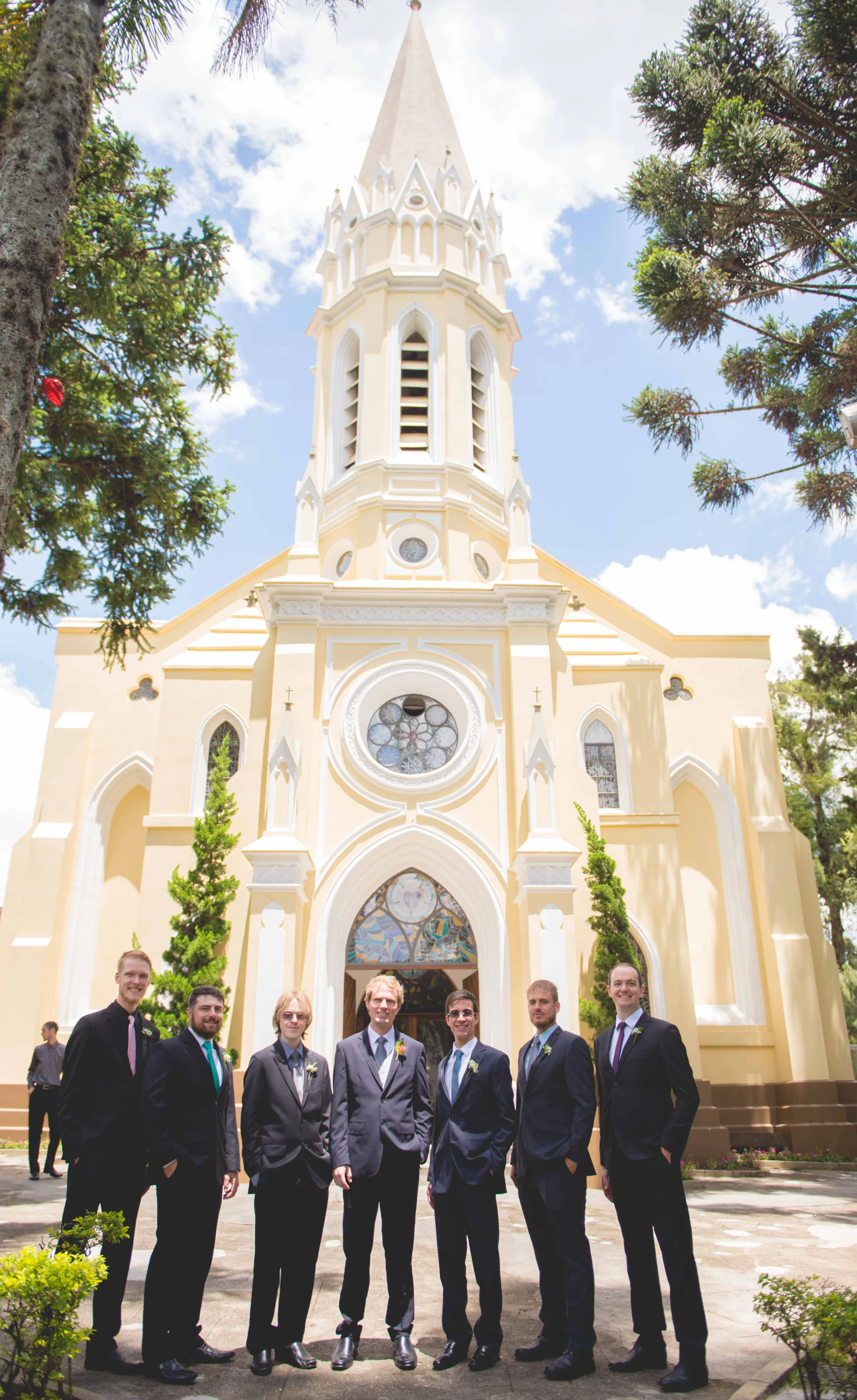 Sete homens de terno enfileirados diante da torre da igreja amarela