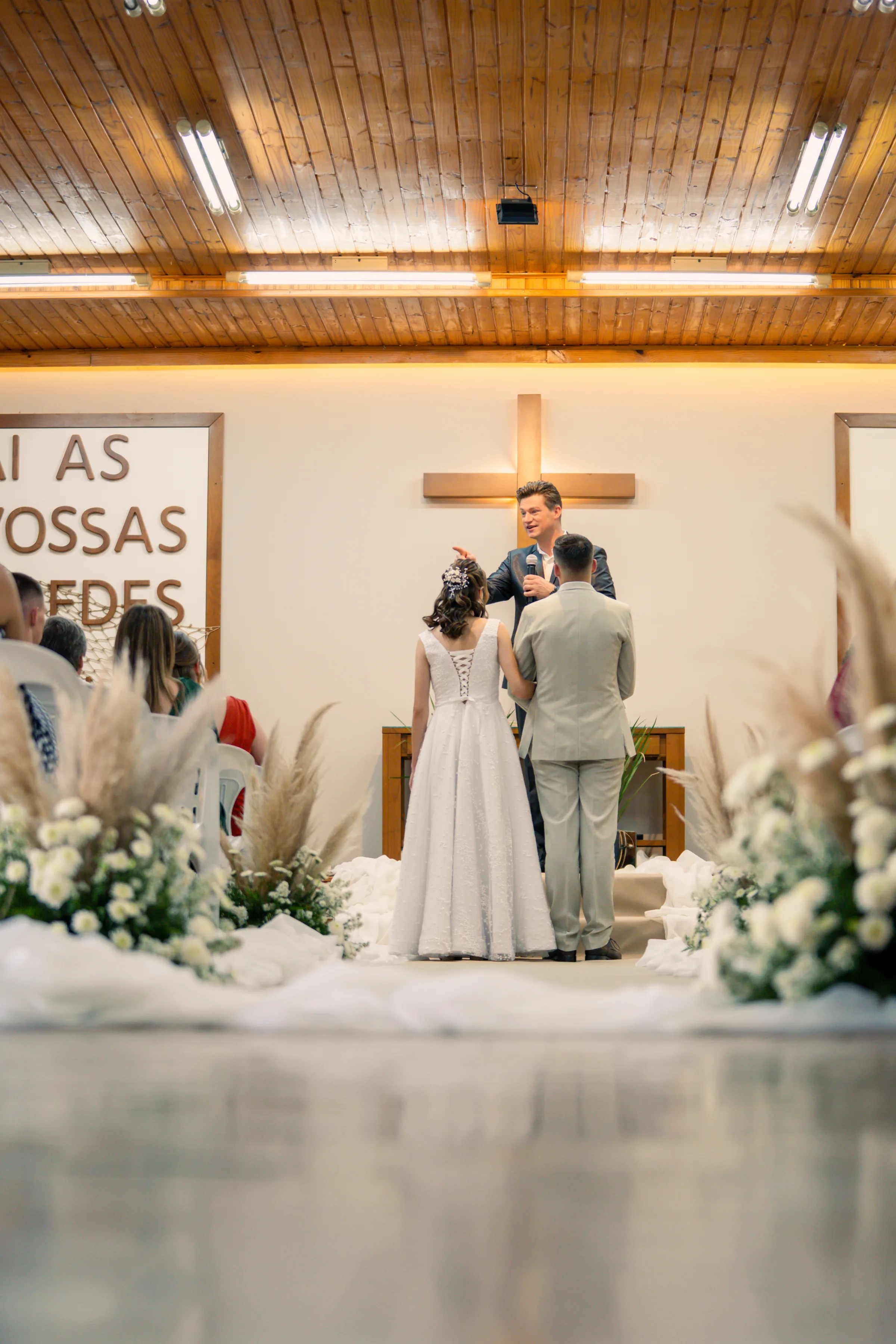 Casal de costas diante do celebrante ao microfone, arranjos de capim dos pampas