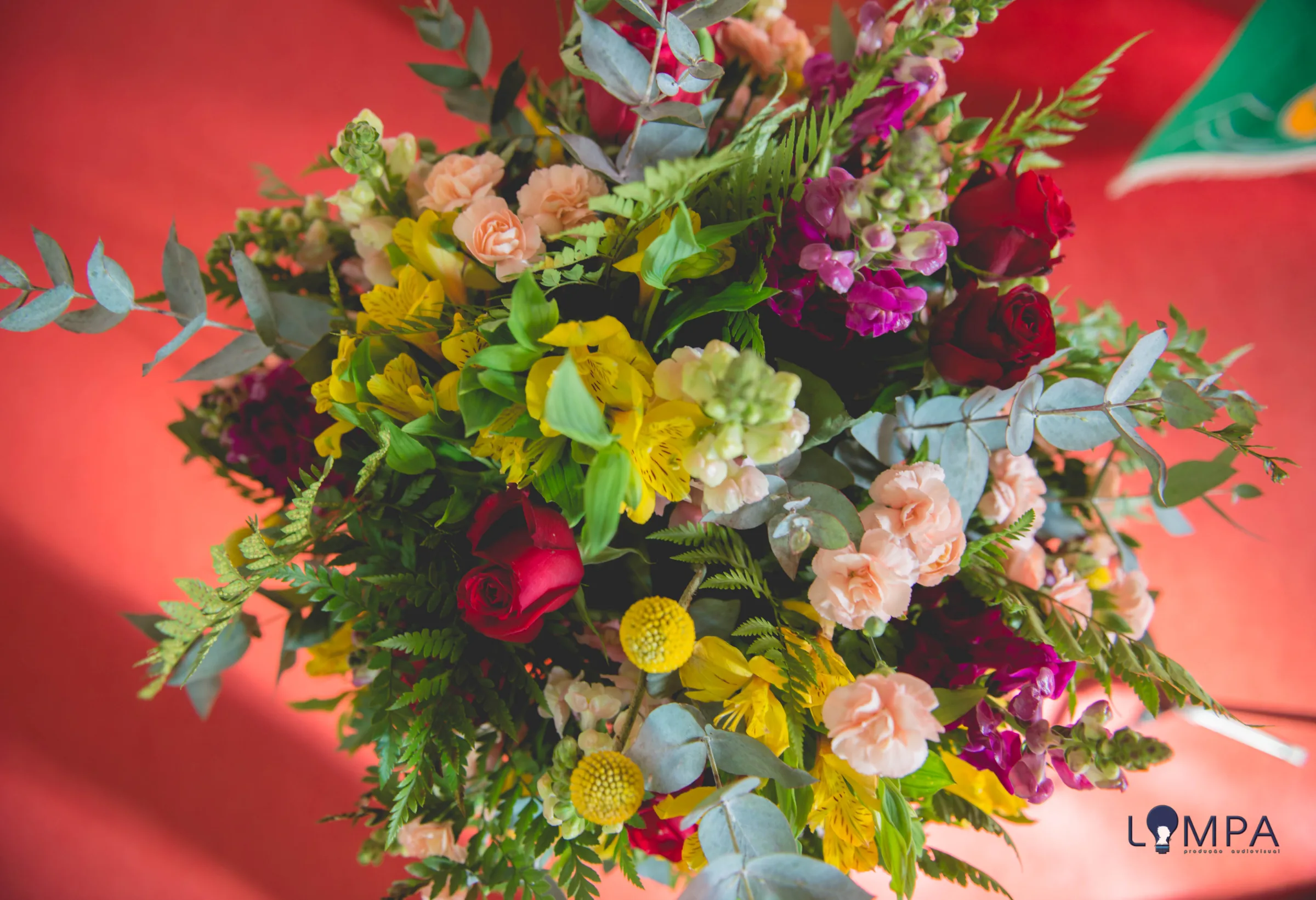 Arranjo visto de cima com rosas, cravos, craspédias e samambaia sobre fundo vermelho