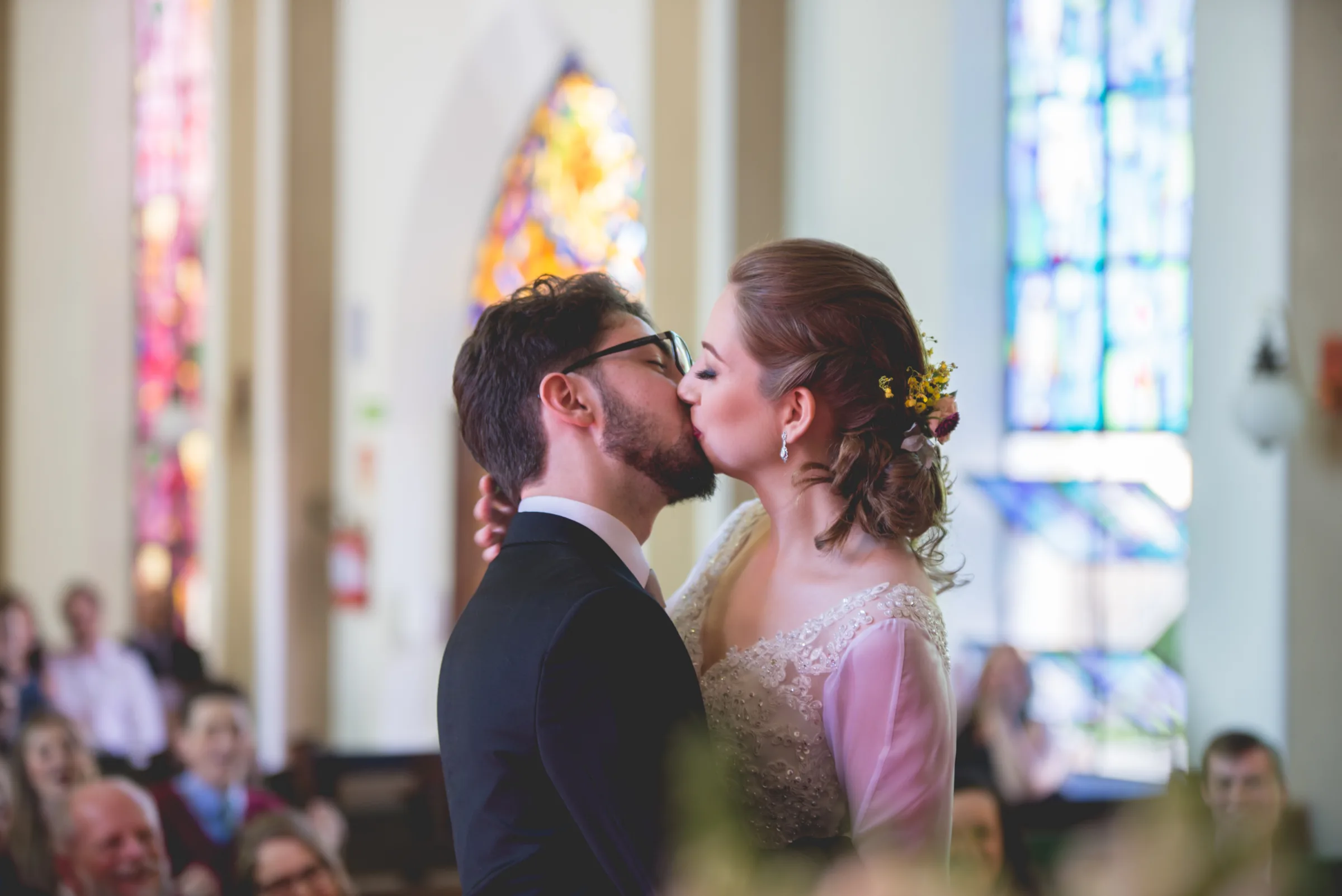 Beijo dos recém-casados diante dos convidados, vitrais coloridos ao fundo