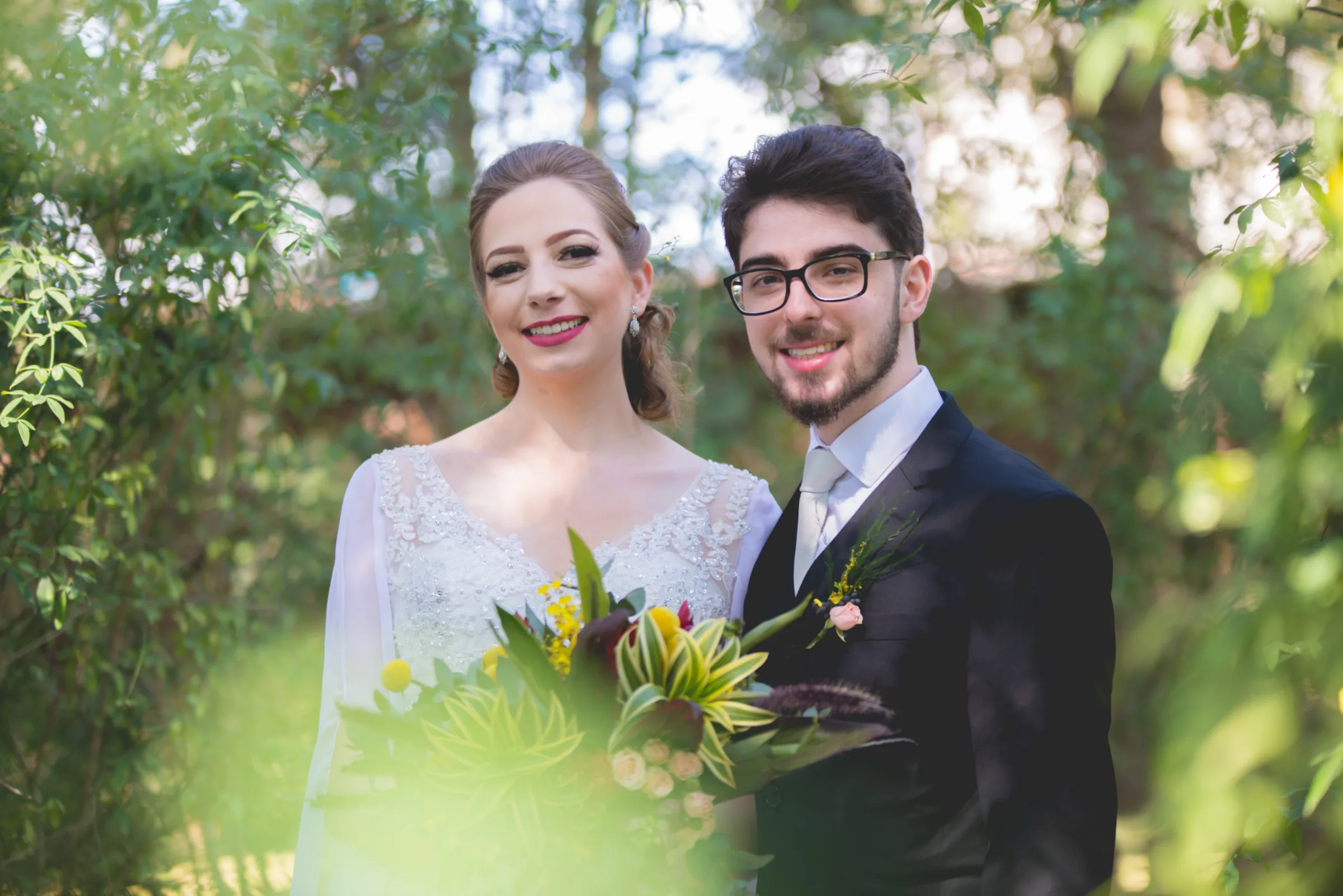 Recém-casados sorrindo para a câmera com buquê tropical, cercados de verde